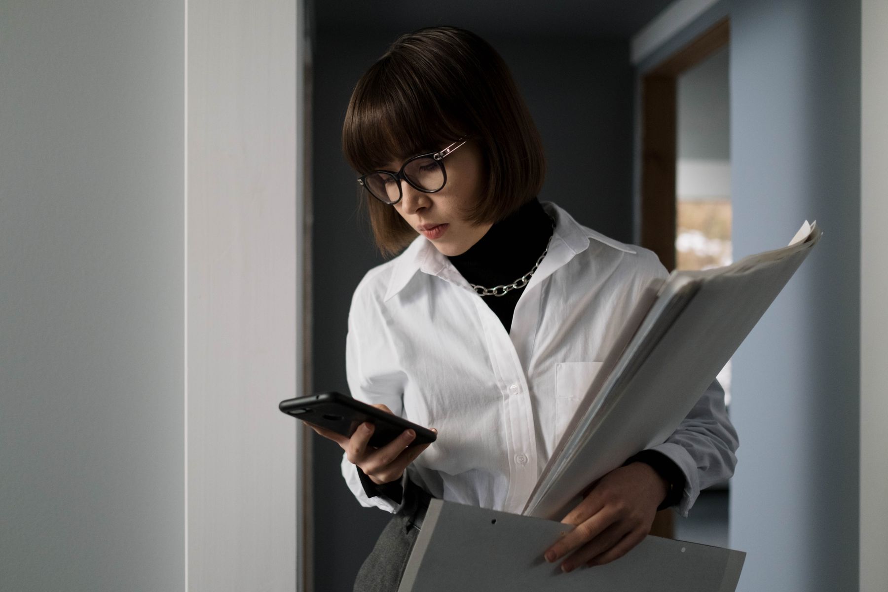 A woman wearing glasses holding her phone in one hand and a folder of paperwork in the other
