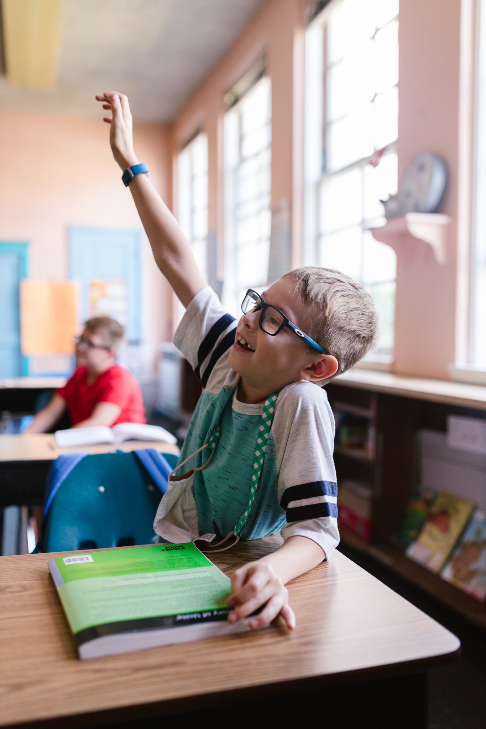 A school-age boy wearing his glasses in class, raising his hand