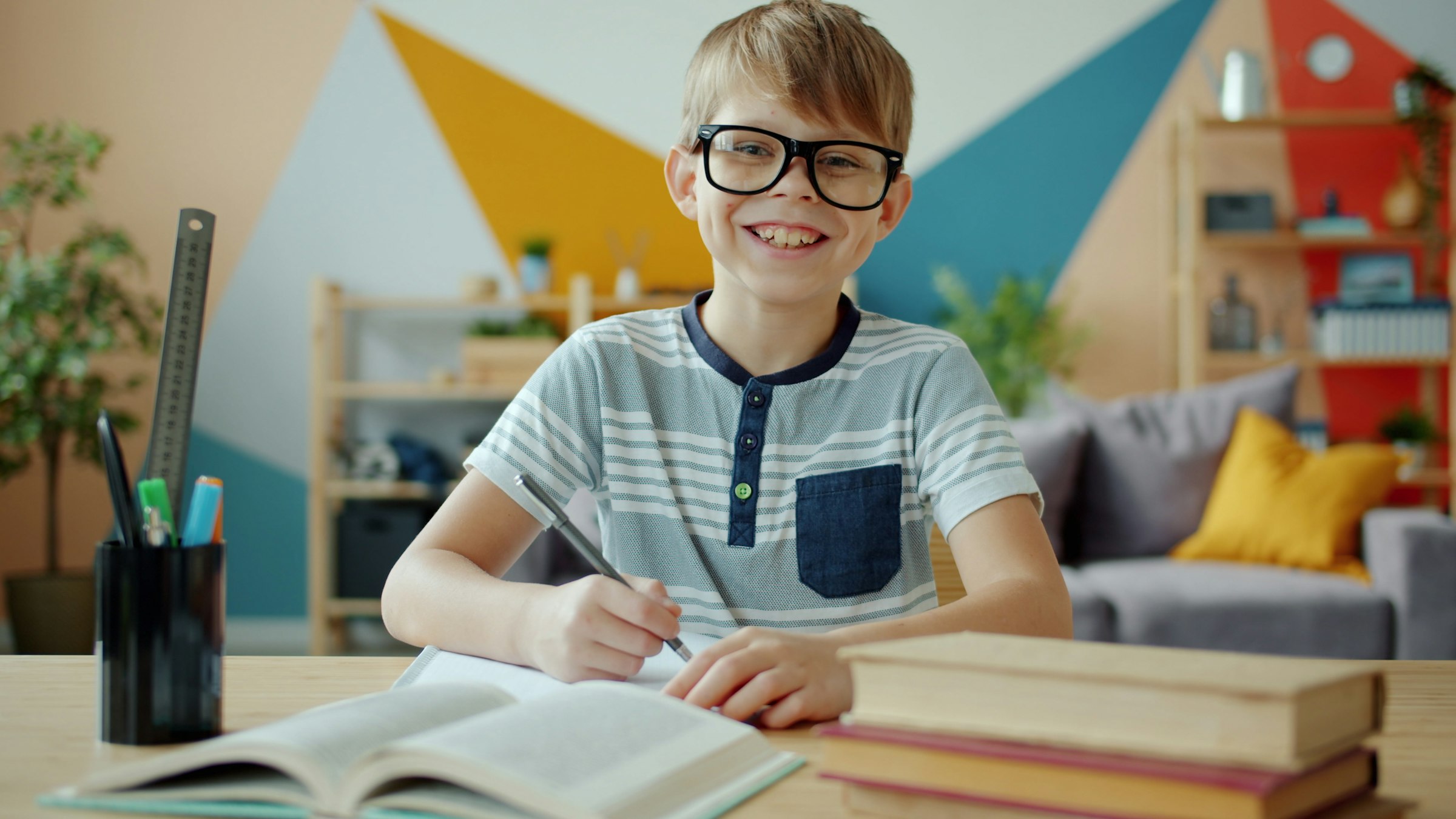 A smiling boy in glasses looking up from his homework at a desk in a bright, colourful room