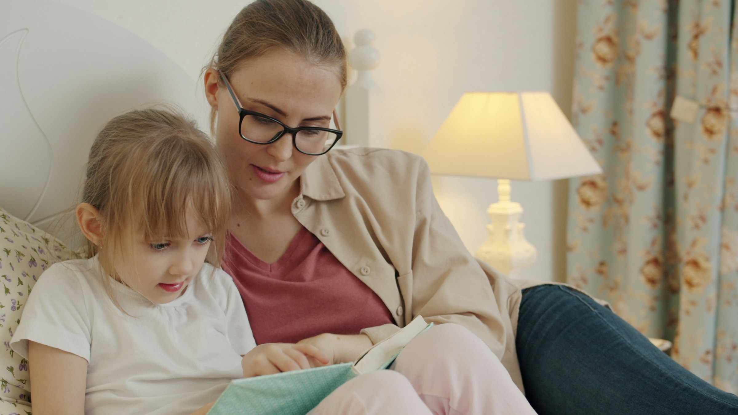 A mother wearing glasses reading a book with her young daughter under a warm lamp